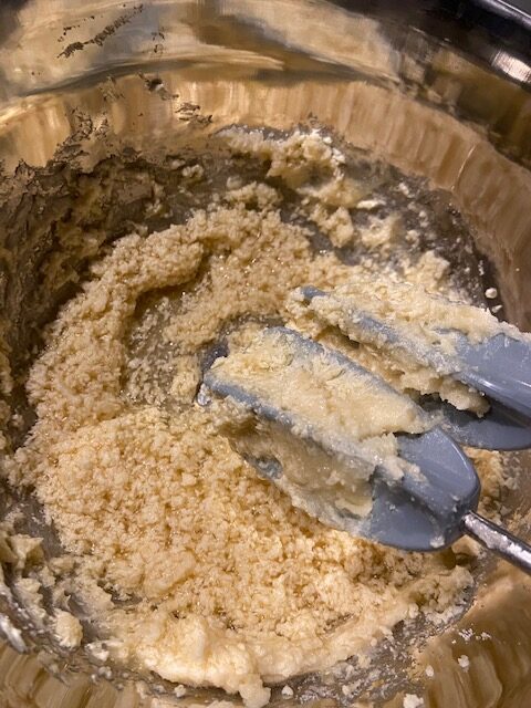 Split butter, sugar, vanilla, and applesauce in stainless steel bowl with the mixer handles in the image. Butter looks chunky like curdles and applesauce is separated from the mix instead of being blended in